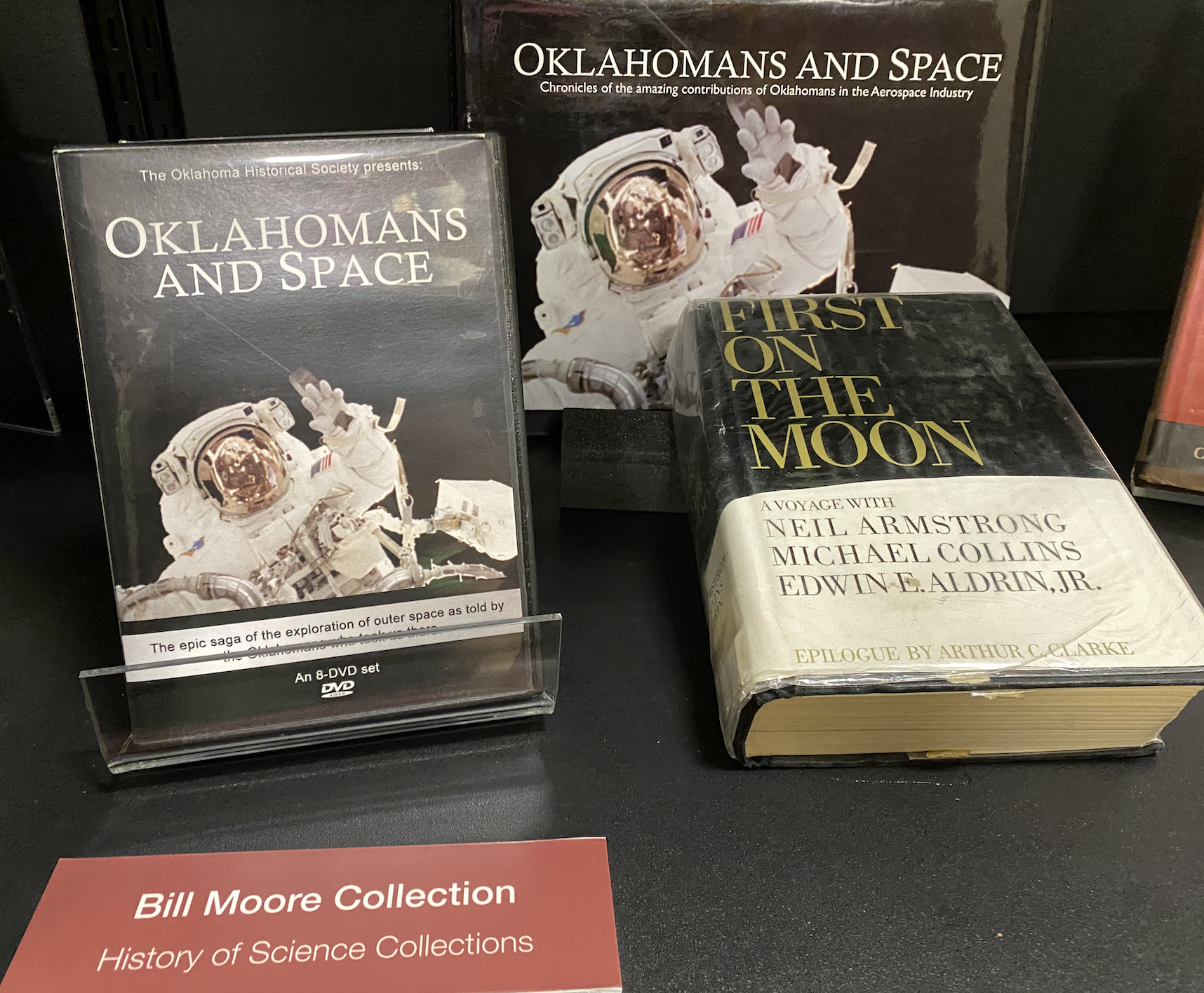 Photo of books on shelf with sign for Bill Moore Collection (includes Oklahomans and Space book & media)