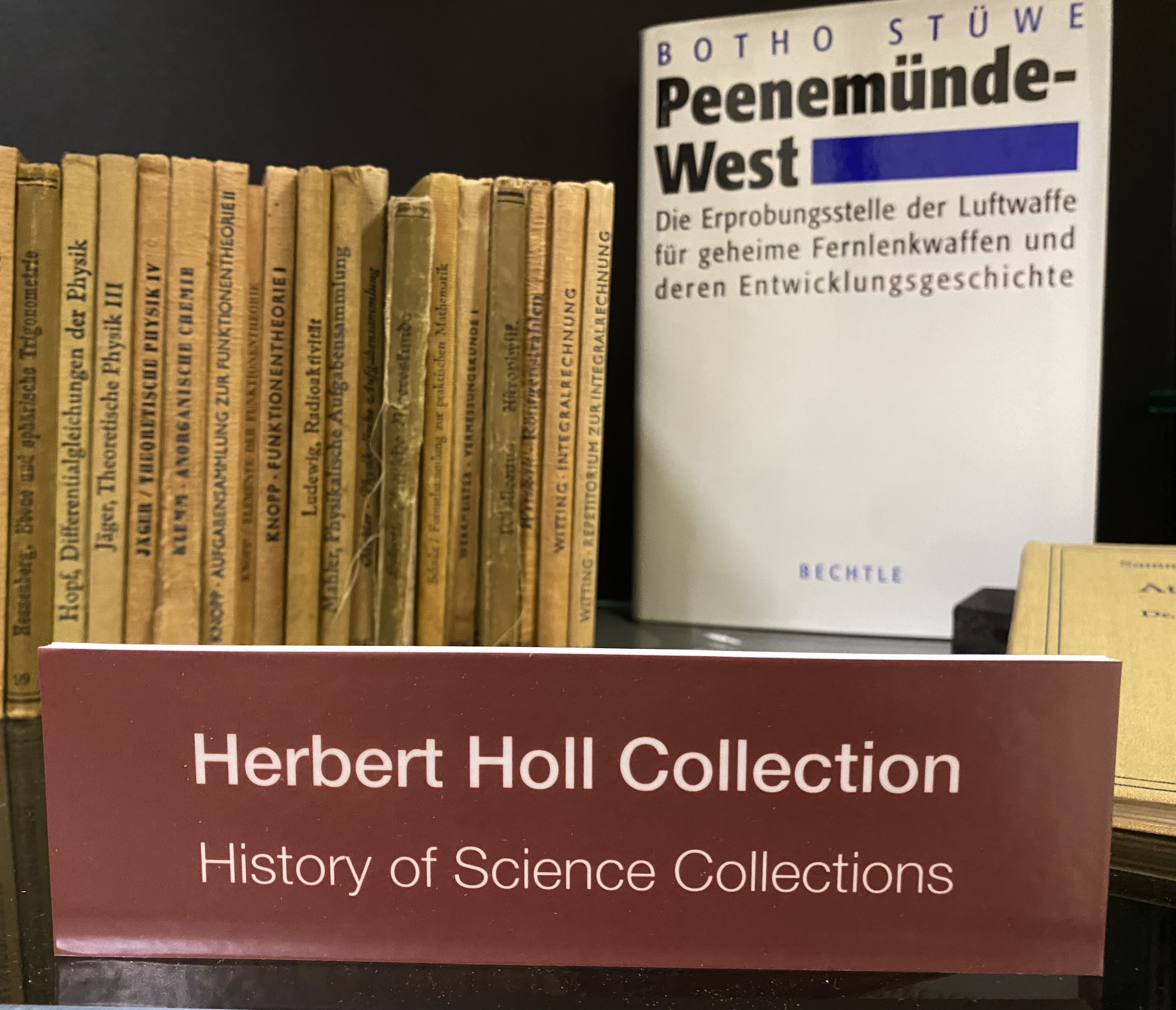 Photo of shelf with small books with spines visible and large book with cover visible; sign says "Herbert Holl Collection"