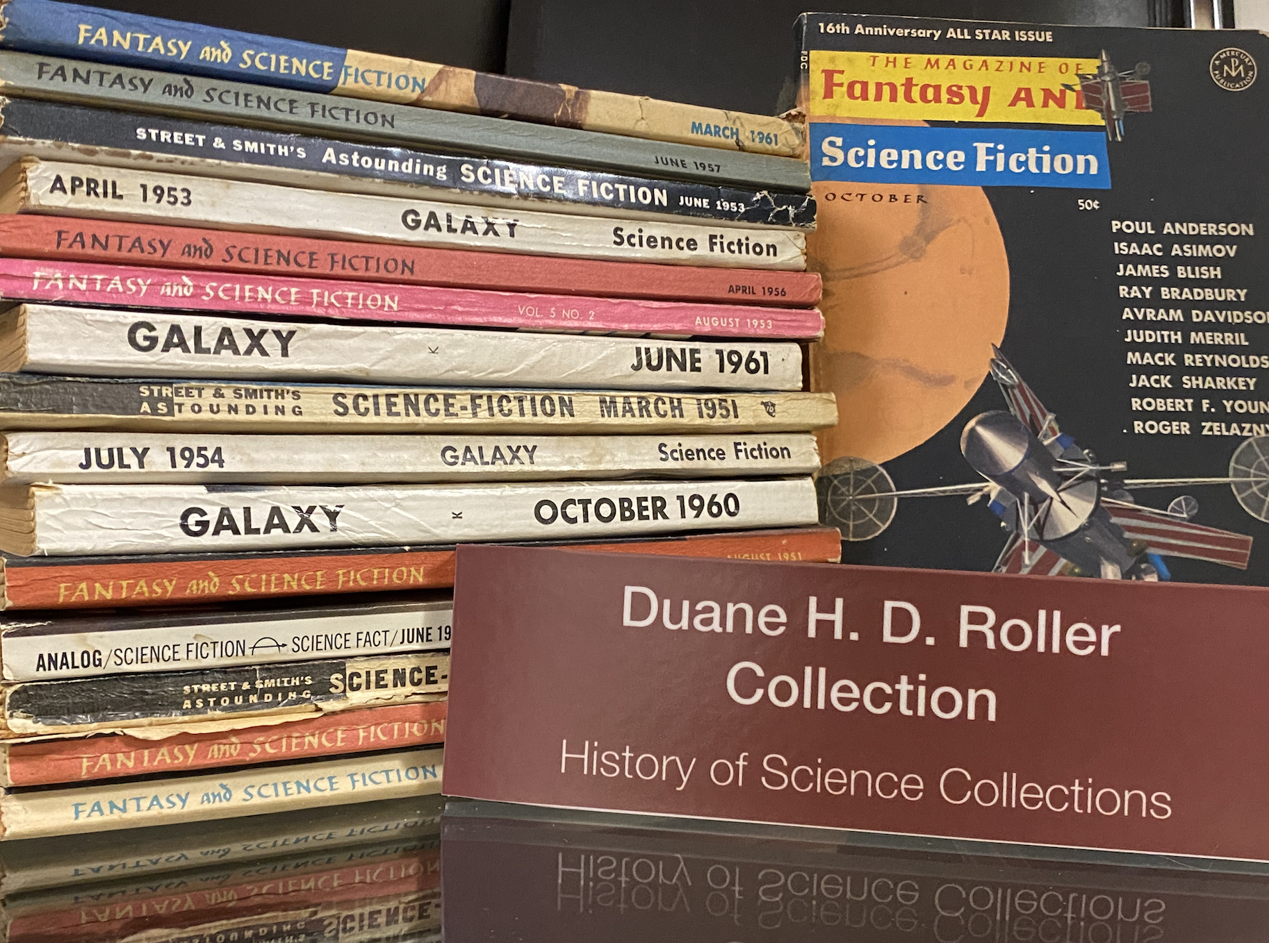 Photo of shelf with stack of pulp science fiction books, and one showing cover; sign says "Duane H. D. Roller Collection"