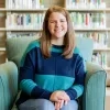 Hallie Macom smiling and sitting in a green chair with books on a shelf behind her.