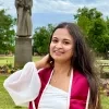 A woman in graduation attire smiles in a park with a statue in the background. She wears a white top and maroon sash.