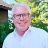 Brian Rossmann is smiling. He has white hair, wears glasses, and is dressed in a white collared shirt. Greenery and a house with a sloped roof are in the background.
