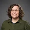 Kristi Wyatt is smiling. She has curly hair, wears glasses and a green button-up shirt, and stands against a neutral gray background.