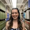Madison is smiling at the camera, standing between shelves of books. She has long brown hair and wears a floral dress.