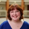 Kennedy Patrick is smiling. She has chin-length red hair and wears glasses. She is wearing a blue V-neck sweater and a gold necklace. She is standing in a softly lit environment with a neutral-colored, blurred building interior in the background.