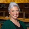 Amanda Palmer is smiling. She has short, light gray hair and is wearing a black V-neck top and a green cardigan. She is in a dim, warmly lit library.