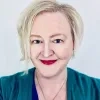 Emrys Moreau has short blonde hair and red lipstick, and she smiles warmly. She wears a teal top, a blue shirt, and a pendant necklace.