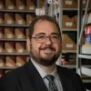 Fred Reiss is smiling. He has short, dark hair and a beard. He is wearing glasses, a gray collared shirt, a patterned black-and-gray tie, and a black suit jacket. He stands in front of shelves filled with organized boxes at the Library Service Center.