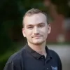 Kyle Brunsmann has short blonde hair and a trimmed goatee. He is wearing a black polo shirt and smiling slightly in an outdoor setting. The background is softly blurred.
