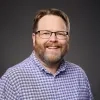 Logan Cox smiles warmly. He has auburn hair and a beard. He wears glasses and a blue-and-purple plaid collared shirt, standing against a gray background.