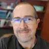 Stewart Brower smiles warmly. He has short hair, a gray beard, and wears glasses. Bookshelves are blurred in the background behind him.