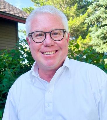 Brian Rossmann is smiling. He has white hair, wears glasses, and is dressed in a white collared shirt. Greenery and a house with a sloped roof are in the background.