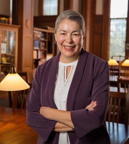 Lina Ortega has gray hair and is smiling towards the camera. She stands confidently in a library, her arms crossed, wearing a purple jacket and white blouse, with warmly lit lamps and bookshelves behind her.